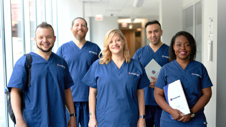 a group of students in blue scrubs smiling and holding program folders for allied health