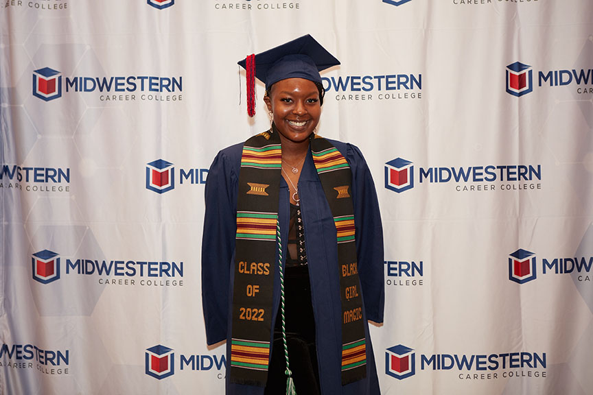 A graduate student in regalia standing in front of a banner with MCC logo as part of the graduating class of 2022.